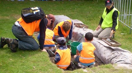Children working in an archaeological trench with a father leaning over the edge looking on. 